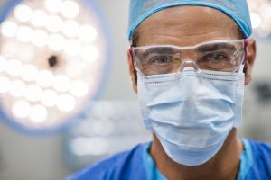 Surgeon with protective mask; Close up face of surgeon man looking at camera with protective mask. Dental assistant with surgical mask and safety glasses in dental clinic. Happy successful surgeon in a operating room.